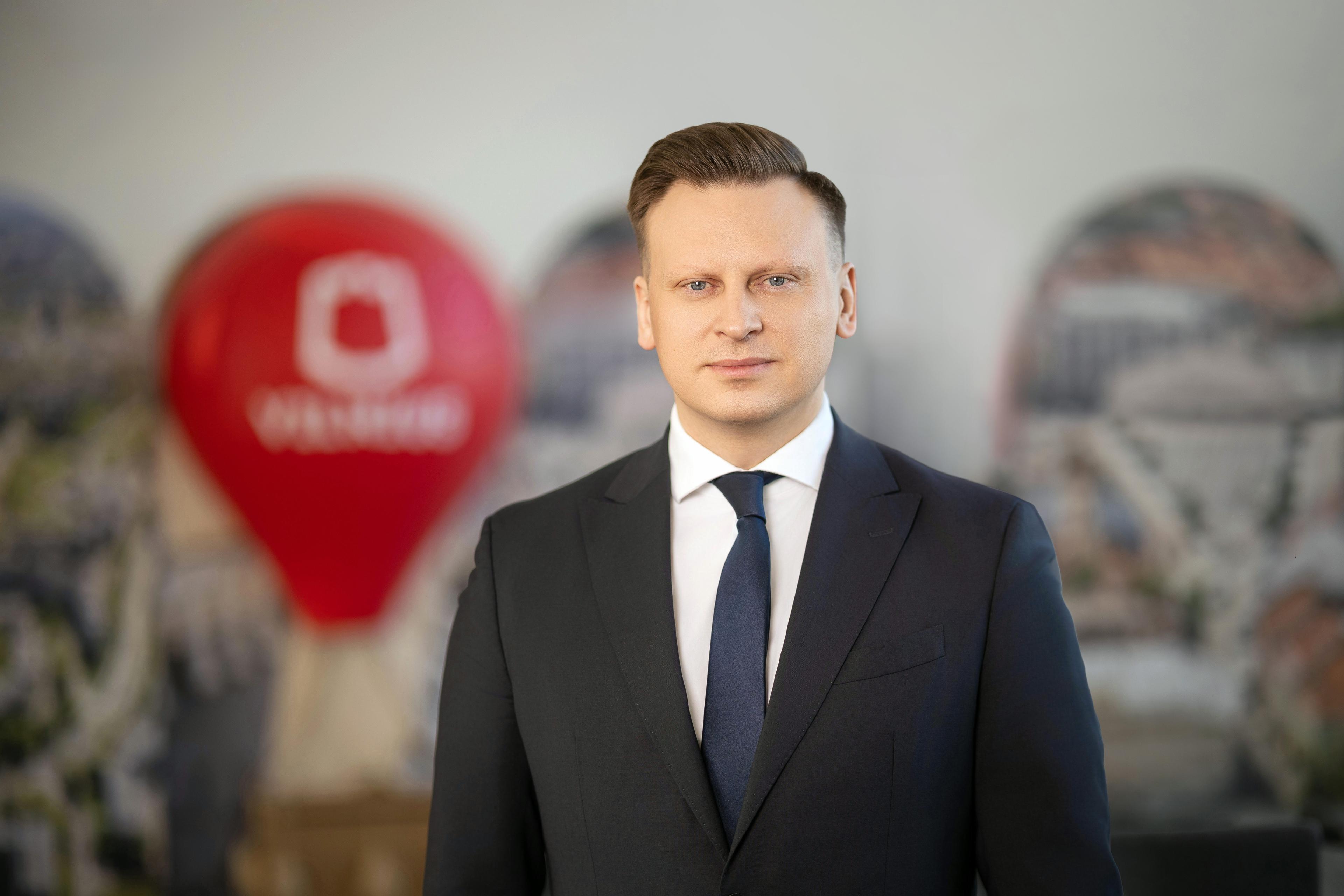 Meras Valdas Benkunskas vilki juodą kostiumą, neutralia išraiška ir švelniu žvilgsniu žvelgia į kamerą, fone matosi Vilniaus oro balionas